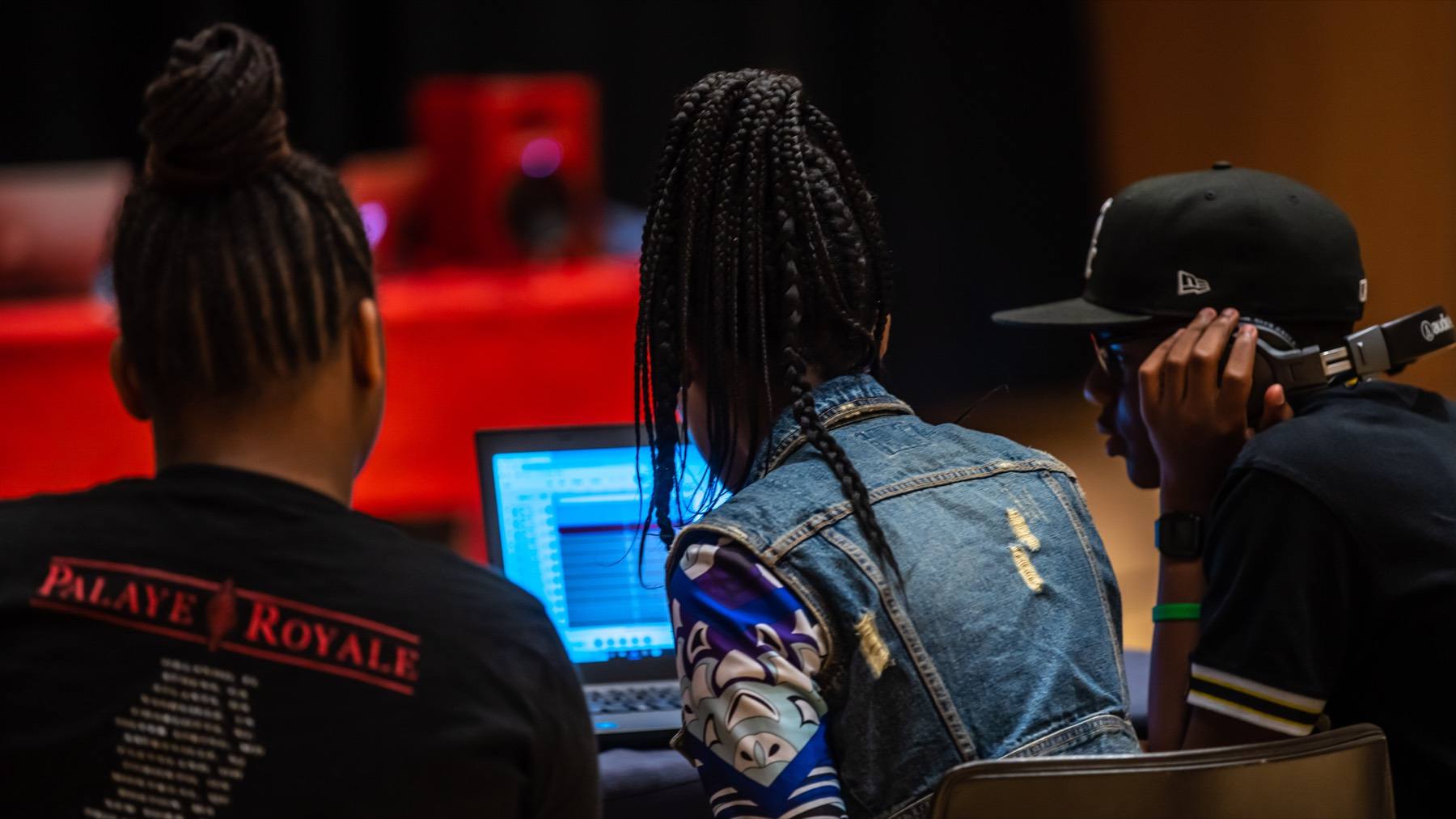 Three students collaborate on music production around a laptop.