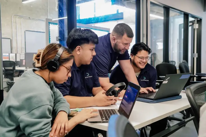 Adult learners and a facilitator collaborate around laptops during a music technology workshop.