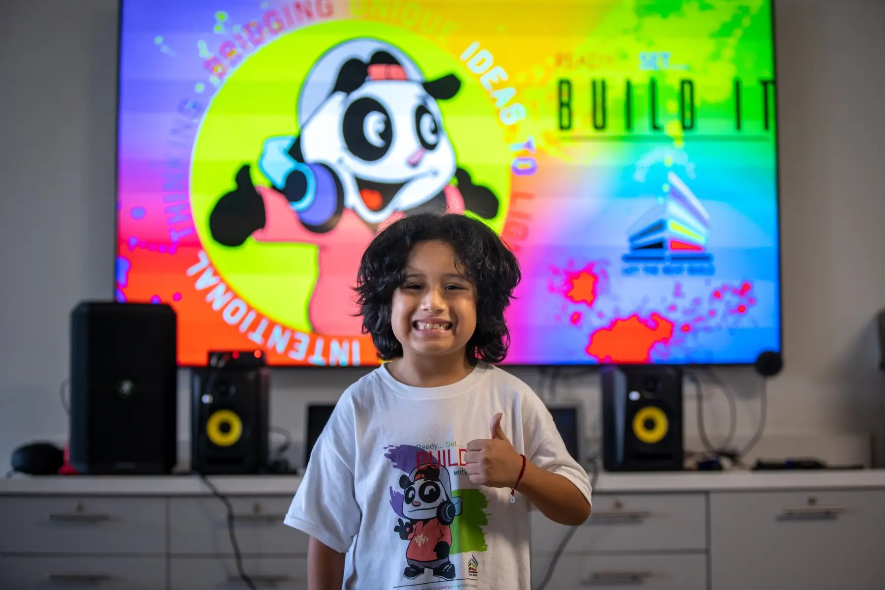 A young LTBB student gives a thumbs-up in front of a BUILD IT studio display.