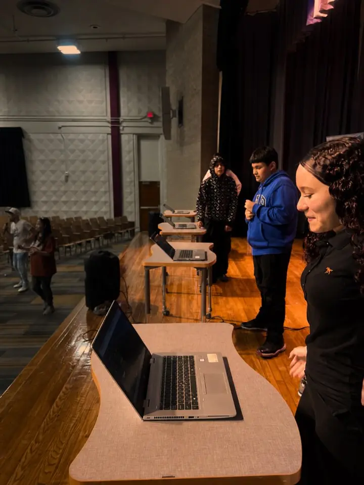 Students stand at laptops on an auditorium stage during a live music creation activity.